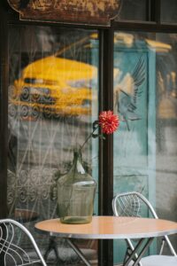 A quaint cafe setting featuring a table with a glass vase and red dahlia, with a city street view.